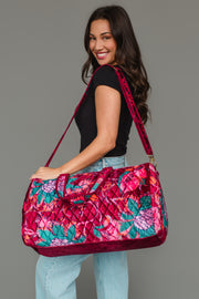 A woman smiles while holding the Panache Apparel Collection Burgundy & Multicolored Floral Duffel, wearing a black top and light blue jeans against a plain gray background.