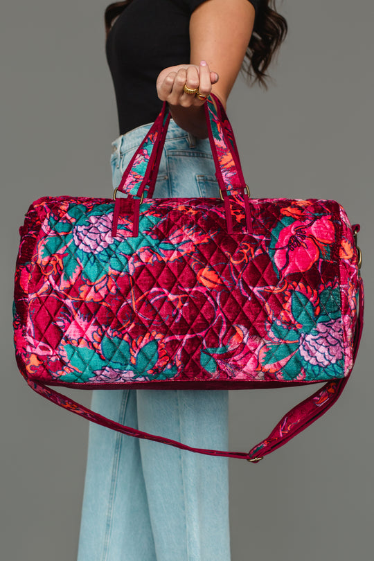 A person wearing a black top and light blue jeans holds the Panache Apparel Collection’s Burgundy & Multicolored Floral Duffel, featuring a large quilted design with vibrant red, pink, and green florals, against a gray background.