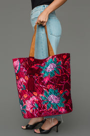 A person in jeans and black heels holds the Panache Apparel Collection Burgundy & Multicolored Floral Tote, a large, colorful quilted bag with floral patterns, brown handles, and a red tassel, against a gray background.