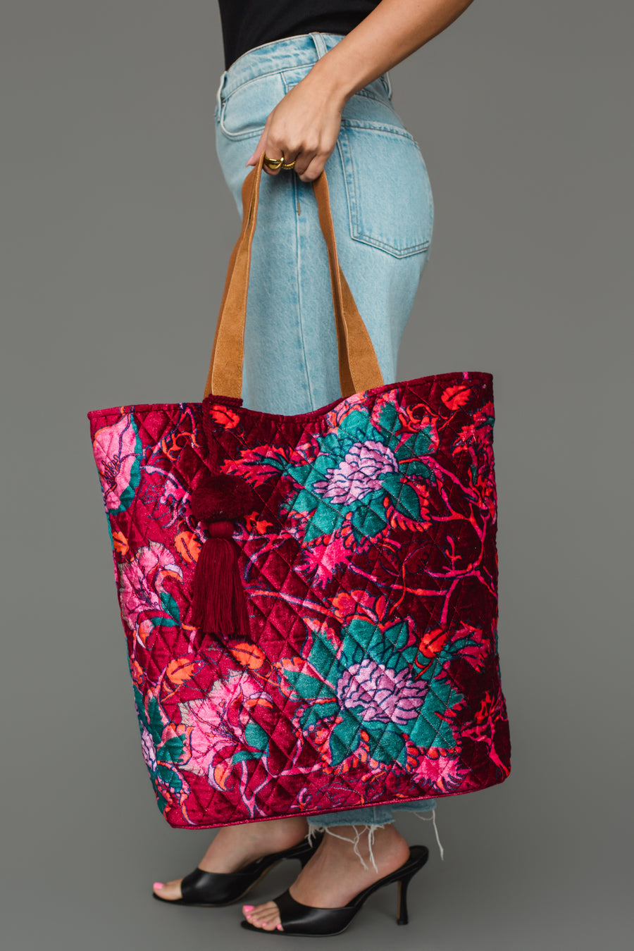 A person in jeans and black heels holds the Panache Apparel Collection Burgundy & Multicolored Floral Tote, a large, colorful quilted bag with floral patterns, brown handles, and a red tassel, against a gray background.