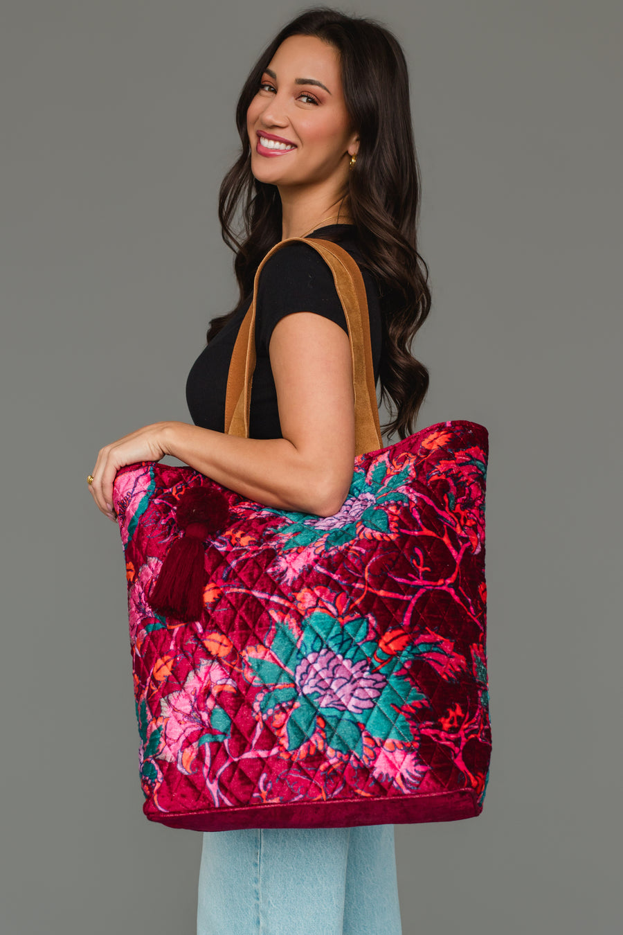 A woman smiles, wearing a black top and jeans, holding the Panache Apparel Collection Burgundy & Multicolored Floral Tote featuring a red, pink, and teal floral pattern with tan straps, against a gray background.