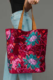 A person holds the Panache Apparel Collection Burgundy & Multicolored Floral Tote, featuring a vibrant red, pink, and teal floral pattern with brown handles and a burgundy tassel. The person is dressed in light blue jeans and a black top.