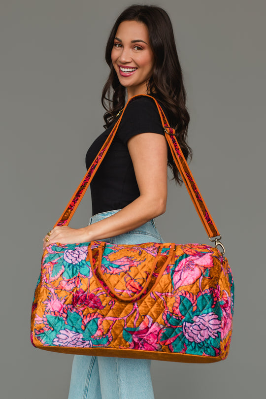 A woman smiles as she poses against a plain gray background, wearing a black top and jeans while holding the Panache Apparel Collection Burnt Orange & Multicolored Floral Duffel, featuring vibrant orange, pink, and teal floral patterns, over her shoulder.