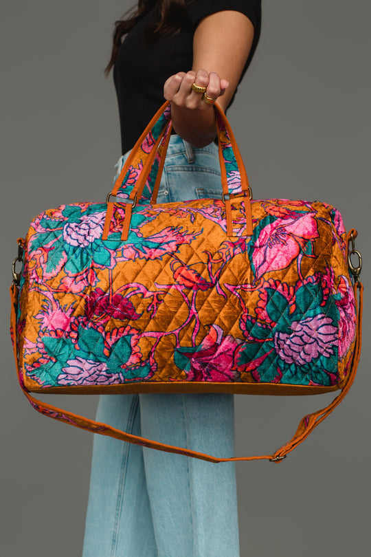 A person in blue jeans and a black top holds the Panache Apparel Collection’s Burnt Orange & Multicolored Floral Duffel, featuring a vibrant floral pattern in pink, teal, and orange, against a plain gray background.