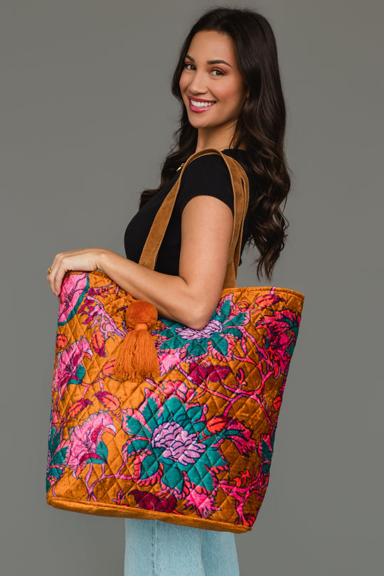 A woman smiles while wearing a black shirt and blue jeans, carrying the Panache Apparel Collection’s Burnt Orange & Multicolored Floral Tote, a large bag featuring vibrant orange, pink, and blue floral patterns with a decorative orange tassel, against a plain gray background.