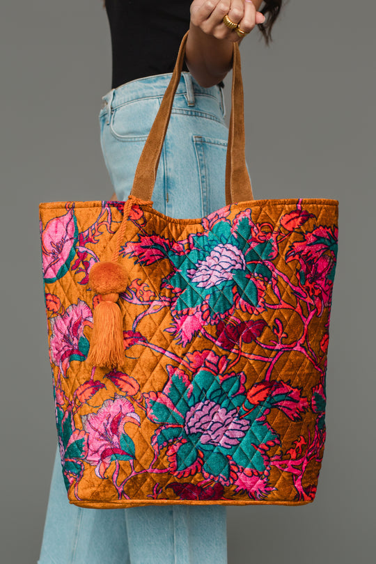A person in jeans holds the Panache Apparel Collection’s Burnt Orange & Multicolored Floral Tote, featuring vibrant orange, pink, and teal patterns, brown handles, and an orange tassel, against a gray background.