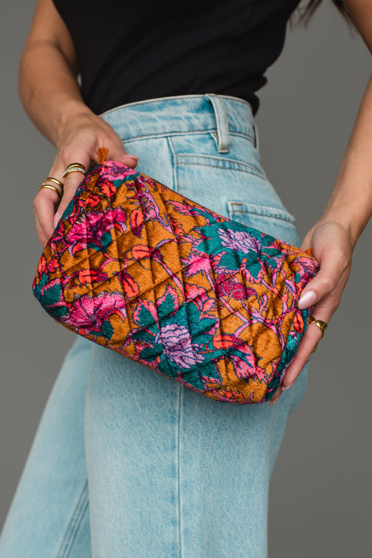 A person in a black top and light blue jeans holds the Burnt Orange & Multicolored Floral Travel Pouch by Panache Apparel Collection, featuring a vibrant quilted pattern with floral designs in pink, teal, orange, and yellow tones.