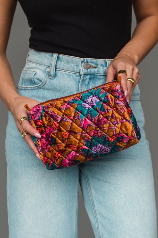 A person in jeans and a black top holds the Panache Apparel Collection Burnt Orange & Multicolored Floral Travel Pouch, featuring a vibrant quilted design with orange, pink, purple, and teal floral patterns.