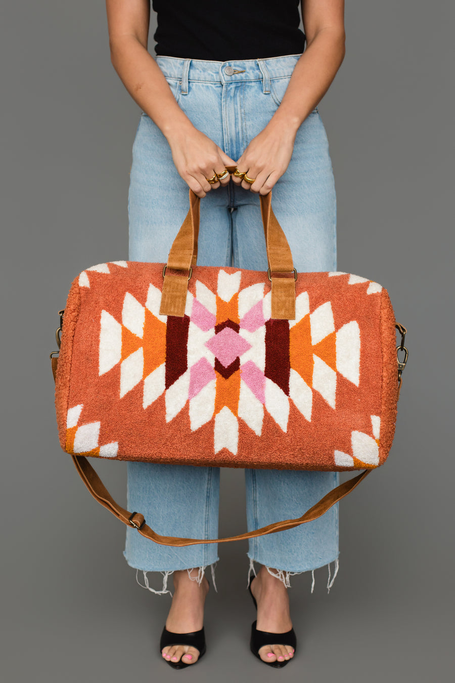 A person in light blue jeans and a black top holds the Panache Apparel Collection’s Rust, Cream & Pink Textured Western Duffel, which features bold geometric designs in orange, white, pink, and brown, while standing against a gray background.