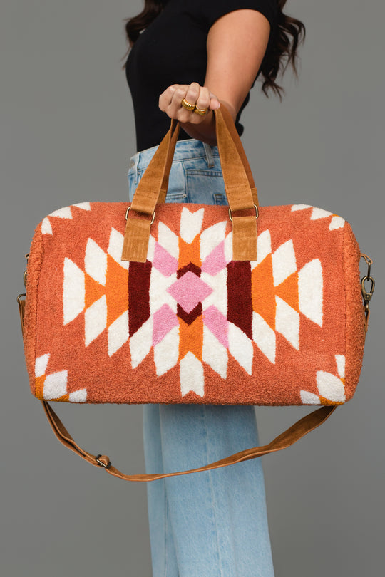 A person holds the Panache Apparel Collection’s Rust, Cream & Pink Textured Western Duffel, which features tan straps and bold geometric designs in orange, white, pink, and red. They are dressed in a black top and light blue jeans.
