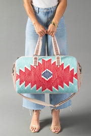 A person in light blue jeans and sandals holds the Panache Apparel Collection Red, Turquoise & Blue Western Duffel, featuring bold geometric patterns, against a gray background.