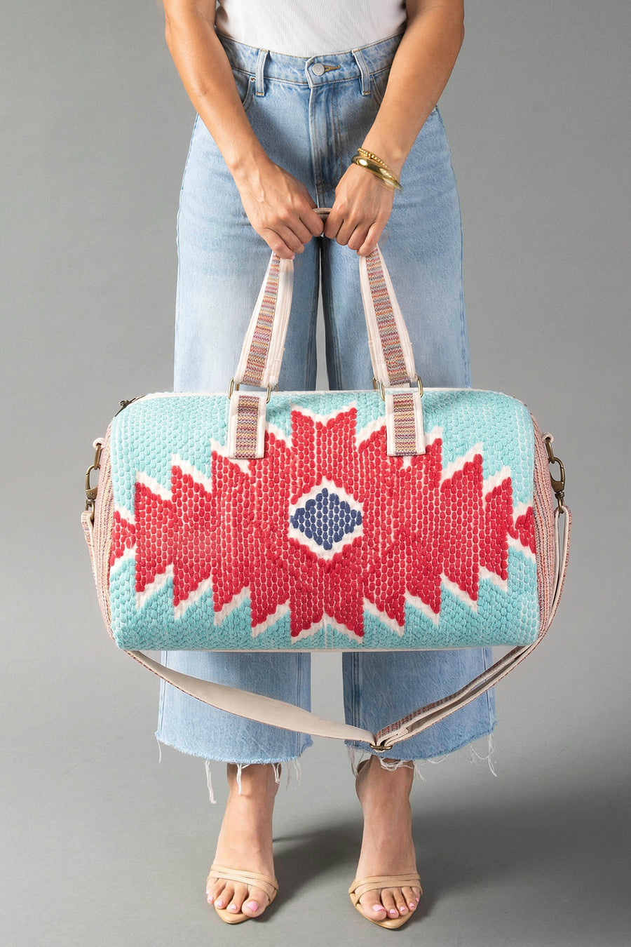 A person in light blue jeans and sandals holds the Panache Apparel Collection Red, Turquoise & Blue Western Duffel, featuring bold geometric patterns, against a gray background.