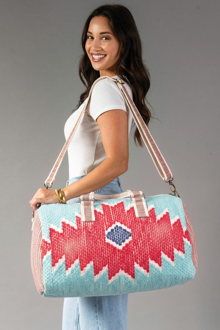 A smiling woman in a white shirt and jeans holds the Panache Apparel Collection Red, Turquoise & Blue Western Duffel, featuring a vibrant geometric pattern and beige straps, against a plain gray background.
