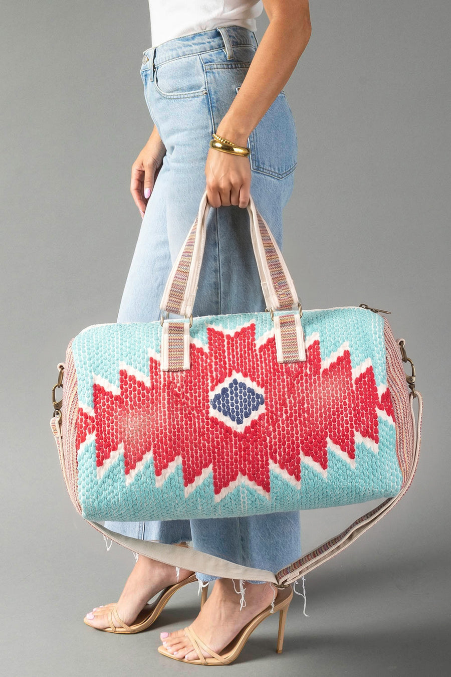 A person in jeans holds the Panache Apparel Collection Red, Turquoise & Blue Western Duffel, which features beige straps and bold geometric patterns, while standing on clear high-heeled shoes.