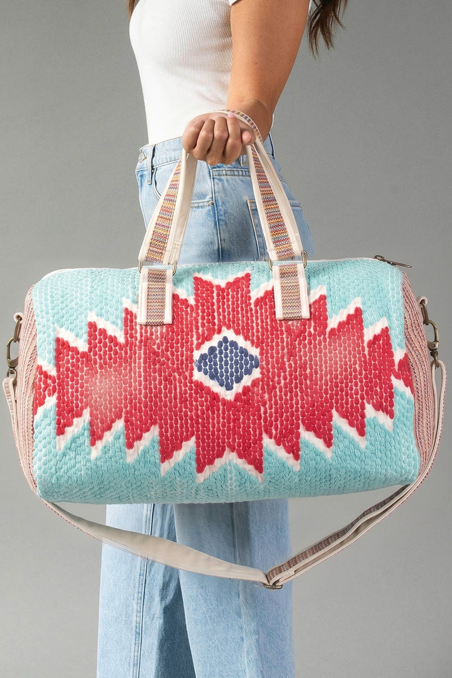 A woman in a white top and blue jeans holds the Red, Turquoise & Blue Western Duffel from Panache Apparel Collection, featuring bold red and blue geometric patterns and beige handles, against a gray background.
