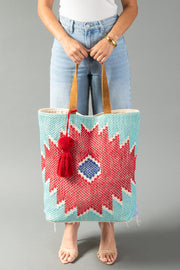 A person in light jeans and sandals holds the Panache Apparel Collection “Red, Turquoise & Blue Western Tote,” featuring tan handles, a red geometric pattern with a blue diamond center, and a red tassel, against a gray background.
