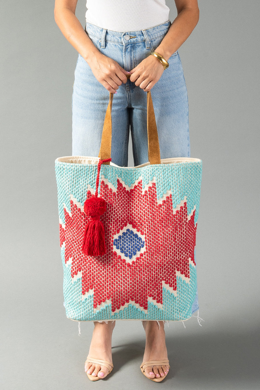 A person in light jeans and sandals holds the Panache Apparel Collection “Red, Turquoise & Blue Western Tote,” featuring tan handles, a red geometric pattern with a blue diamond center, and a red tassel, against a gray background.