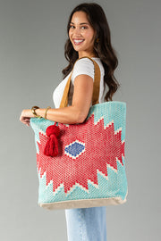A woman in a white top and jeans smiles while holding the Panache Apparel Collection’s Red, Turquoise & Blue Western Tote, a large woven bag featuring red, blue, and white geometric patterns with a red tassel, against a plain gray background.