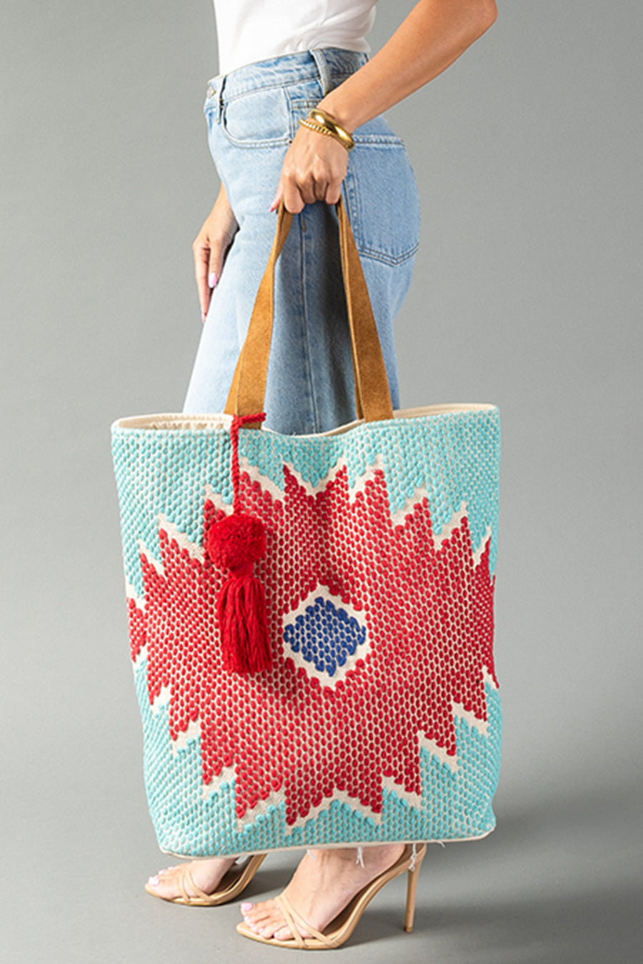 A person in jeans and heels holds the Panache Apparel Collection’s Red, Turquoise & Blue Western Tote, featuring a bold geometric pattern in blue, red, and white with a red tassel and tan shoulder straps.
