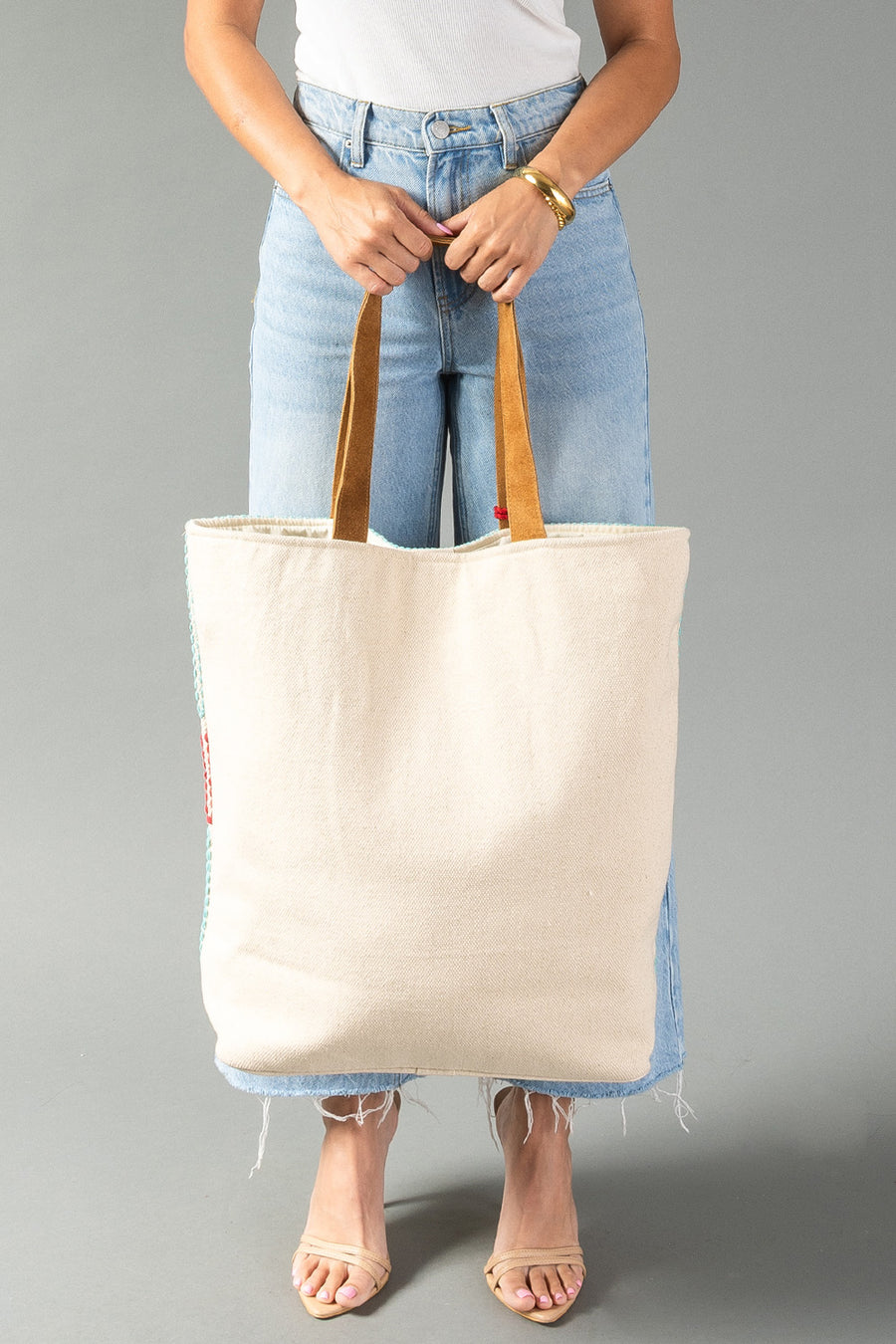 A person in a white top and blue jeans holds the Red, Turquoise & Blue Western Tote from Panache Apparel Collection. Their feet are in beige sandals and the background is plain gray.