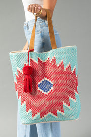 A person wearing light blue jeans holds the Panache Apparel Collection Red, Turquoise & Blue Western Tote, featuring tan handles, a red and blue geometric pattern, and a red tassel, against a plain gray background.