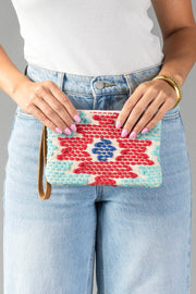 A person in light blue jeans and a white top holds the Panache Apparel Collection’s Red, Turquoise & Blue Western Wristlet, a colorful woven clutch featuring red, blue, and turquoise patterns. They have pink nails and wear gold bangles.