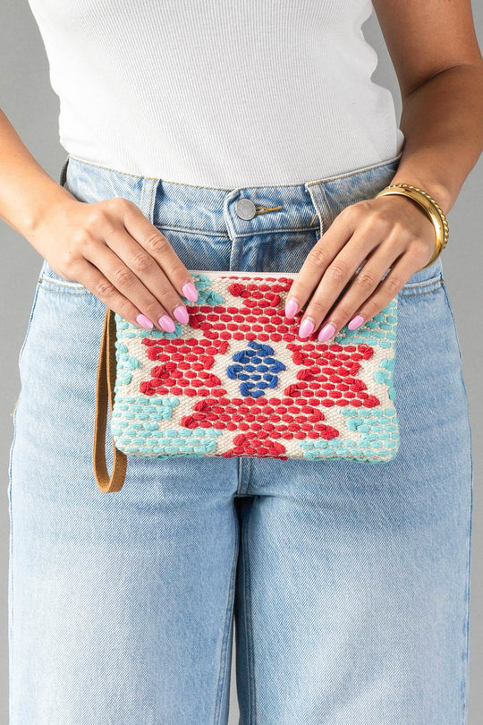 A person in light blue jeans and a white top holds the Panache Apparel Collection’s Red, Turquoise & Blue Western Wristlet, a colorful woven clutch featuring red, blue, and turquoise patterns. They have pink nails and wear gold bangles.