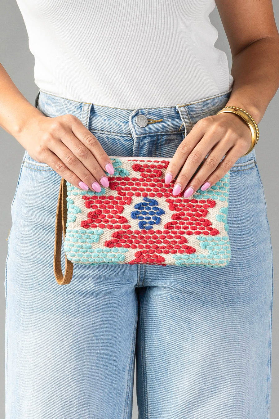 A person in light blue jeans and a white top holds the Panache Apparel Collection’s Red, Turquoise & Blue Western Wristlet, a colorful woven clutch featuring red, blue, and turquoise patterns. They have pink nails and wear gold bangles.