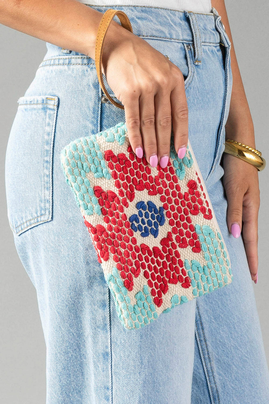 A person wearing blue jeans holds the Red, Turquoise & Blue Western Wristlet by Panache Apparel Collection, a small clutch featuring a vibrant geometric pattern in red, turquoise, and blue, complete with a tan strap.