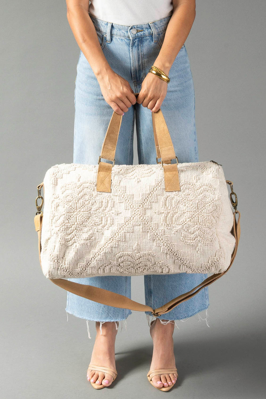 A person wearing light blue jeans is holding the Cream Vintage Inspired Textured Duffel from Panache Apparel Collection, a large cream-colored bag featuring a subtle patterned texture, tan straps, and a detachable shoulder strap. The person also accessorizes with sandals and a gold bracelet.