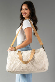 A smiling woman with long dark hair, dressed in a white shirt and jeans, stands against a plain gray background holding the Panache Apparel Collection’s Cream Vintage Inspired Textured Duffel, featuring tan straps, over her shoulder.