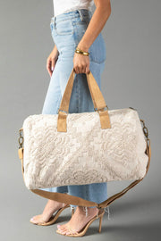 A person in jeans and heels showcases the Panache Apparel Collection Cream Vintage Inspired Textured Duffel, featuring tan handles and a shoulder strap, against a gray background.