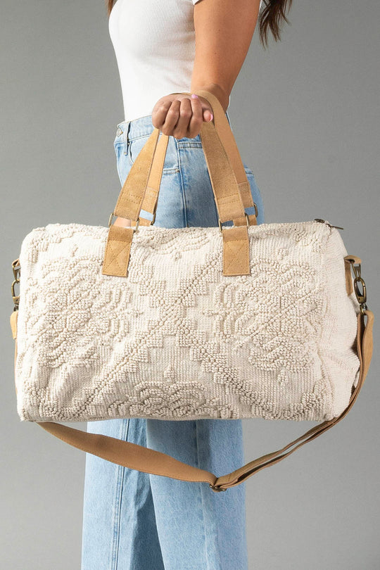 A person wearing a white tank top and blue jeans holds the Panache Apparel Collection’s Cream Vintage Inspired Textured Duffel, featuring tan leather handles and a matching shoulder strap, against a plain gray background.