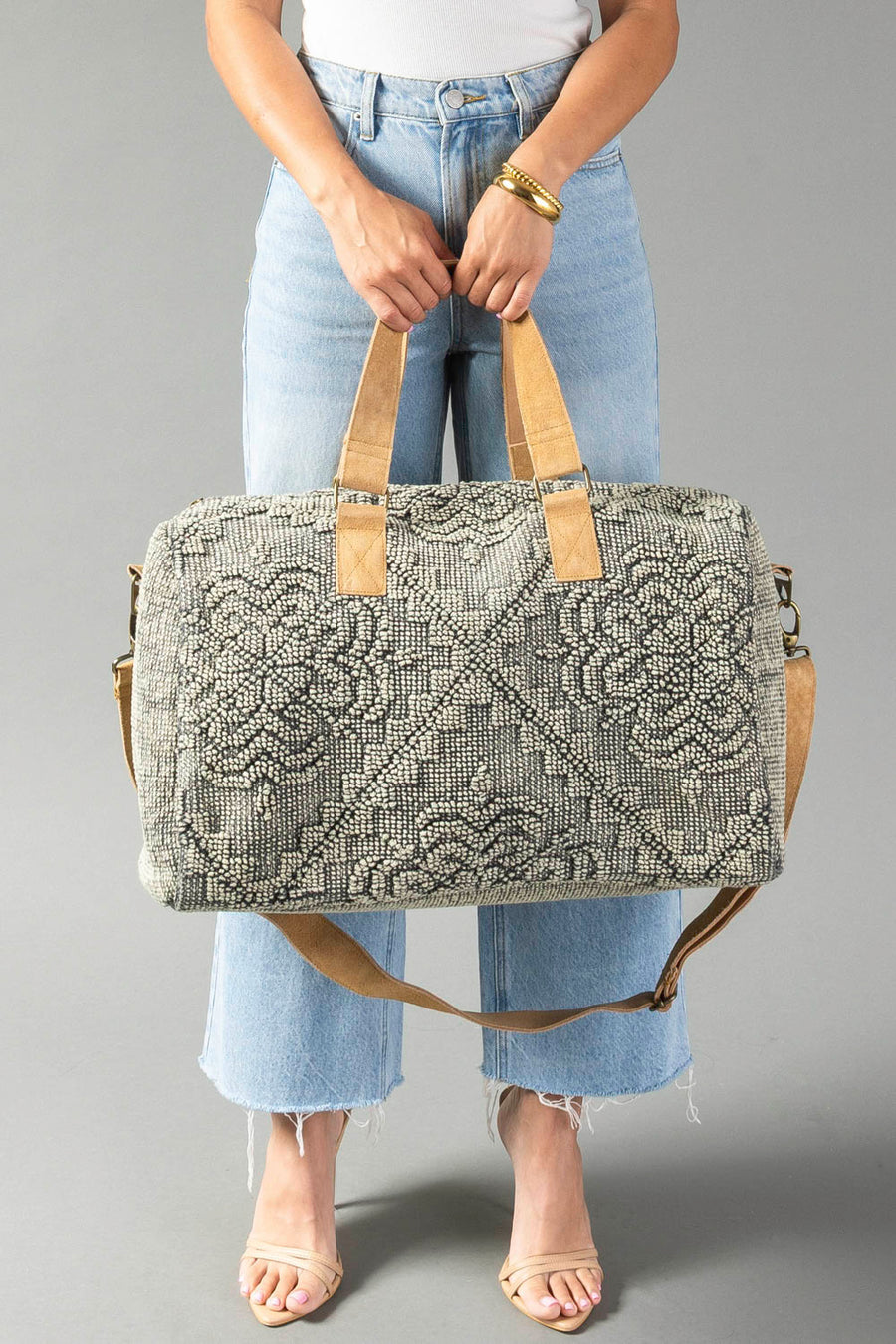 Wearing light jeans and a white top, a person holds the Panache Apparel Collection Gray Vintage Inspired Textured Duffel with tan handles and a detachable strap, standing against a plain gray background.