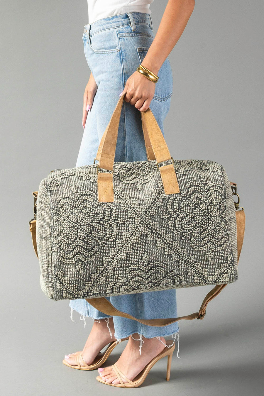 A woman in light blue jeans and heeled sandals holds the Gray Vintage Inspired Textured Duffel by Panache Apparel Collection, featuring tan handles and a matching shoulder strap, against a plain gray background.