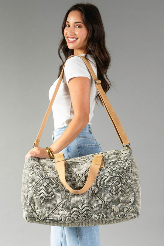 A smiling woman with long brown hair, in a white t-shirt and light blue jeans, carries the Gray Vintage Inspired Textured Duffel by Panache Apparel Collection over her shoulder against a plain gray background.