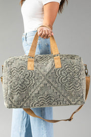 A person in a white shirt and blue jeans holds the Gray Vintage Inspired Textured Duffel by Panache Apparel Collection, featuring gray patterns, tan handles, and a matching shoulder strap against a plain background.