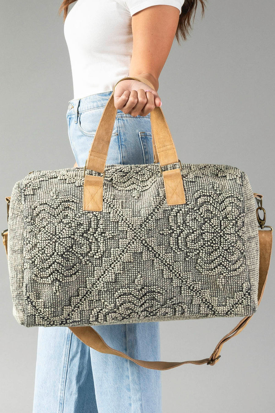 A person in a white shirt and blue jeans holds the Gray Vintage Inspired Textured Duffel by Panache Apparel Collection, featuring gray patterns, tan handles, and a matching shoulder strap against a plain background.