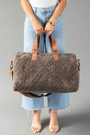 A person wearing light blue jeans and a white top holds the Brown Vintage Inspired Textured Duffel from Panache Apparel Collection, featuring brown handles and strap, standing against a gray background.