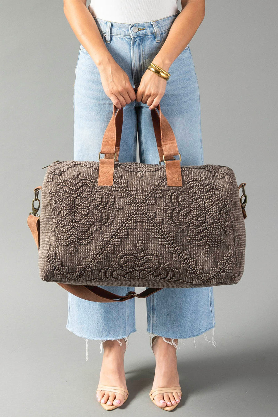 A person wearing light blue jeans and a white top holds the Brown Vintage Inspired Textured Duffel from Panache Apparel Collection, featuring brown handles and strap, standing against a gray background.