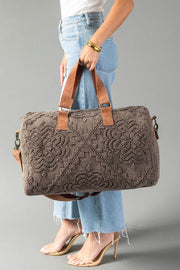 A woman in a white shirt and light blue jeans carries the Panache Apparel Collection Brown Vintage Inspired Textured Duffel, featuring a floral pattern, leather handles, and a detachable shoulder strap. She accessorizes with heeled sandals and gold bangles.