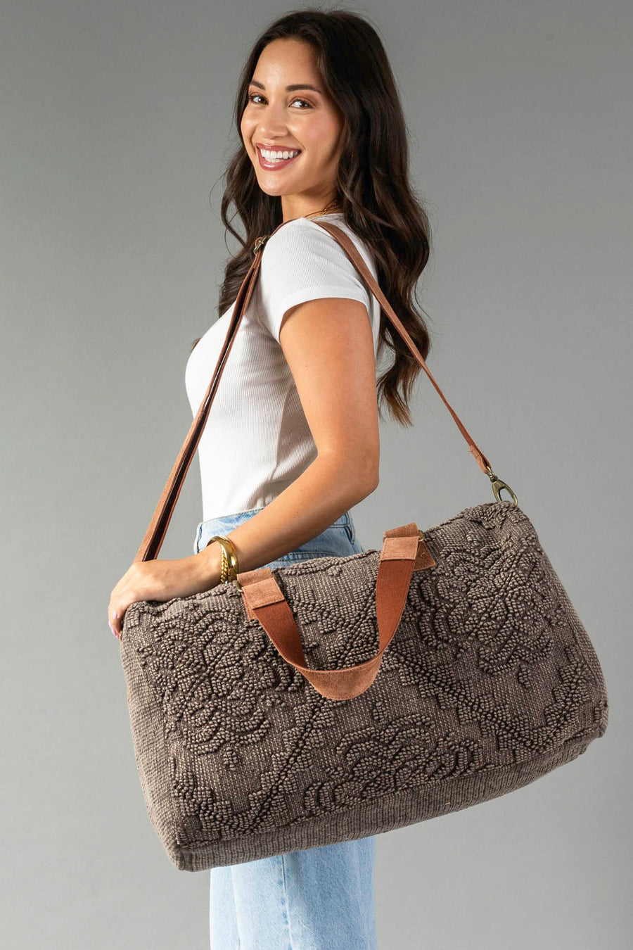 A woman smiles while wearing a white t-shirt and jeans, carrying the Panache Apparel Collection’s Brown Vintage Inspired Textured Duffel, featuring tan handles and a long shoulder strap, against a plain gray background.