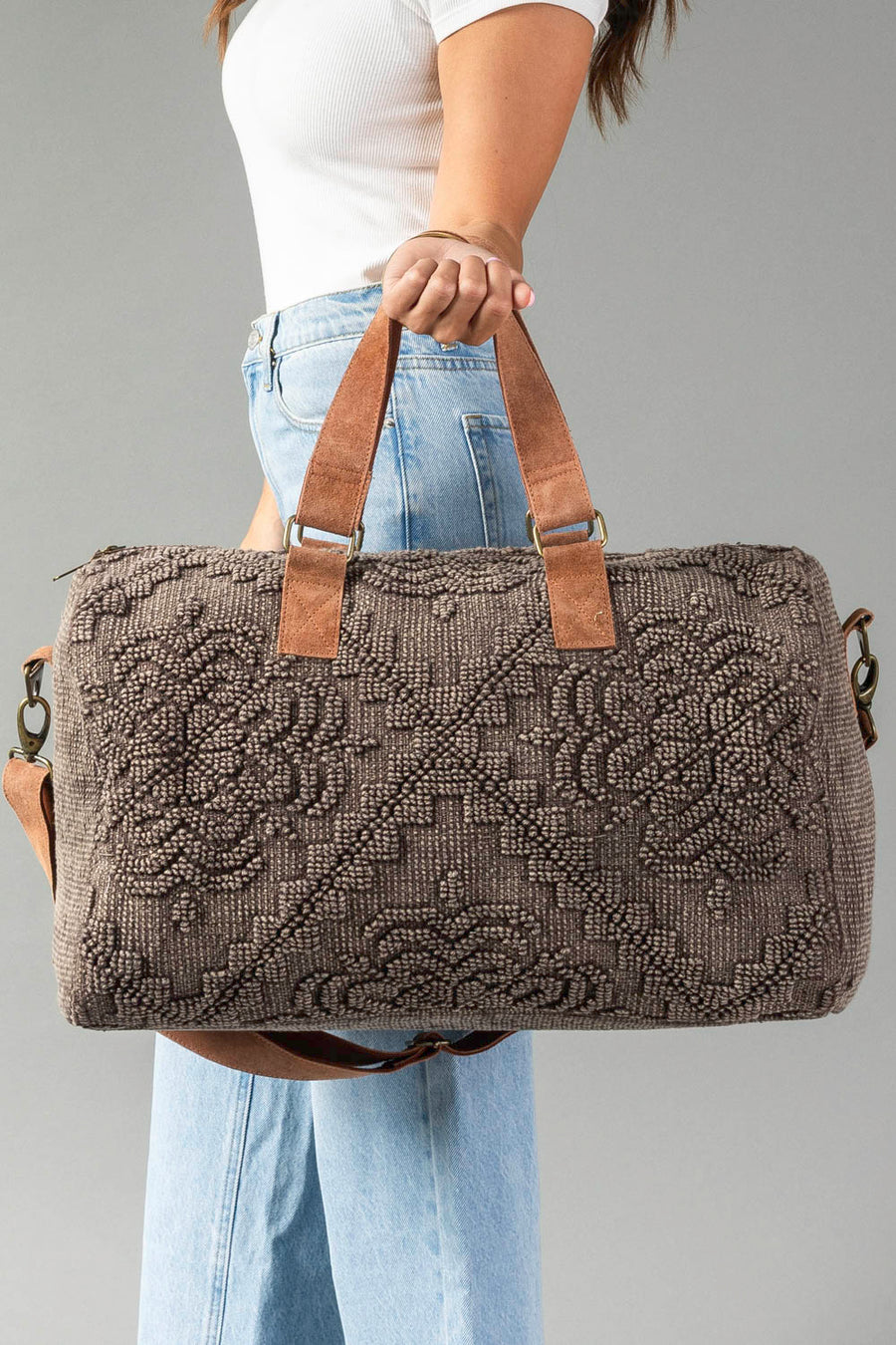 A woman in a white top and blue jeans holds the Panache Apparel Collection Brown Vintage Inspired Textured Duffel against a plain gray background.