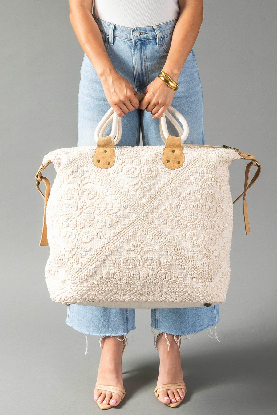 A person wearing light blue jeans and a white top holds the Cream Vintage Inspired Textured Tote by Panache Apparel Collection, which features tan leather handles. They are also wearing beige sandals and a gold bracelet.