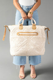 A person in light jeans and sandals holds the Panache Apparel Collection Cream Vintage Inspired Textured Tote, featuring tan handles and a front zipper pocket, against a gray background.