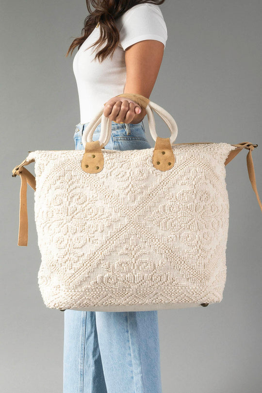 A woman in a white top and blue jeans holds the Panache Apparel Collection’s Cream Vintage Inspired Textured Tote, featuring short handles and tan accents, against a plain gray background.