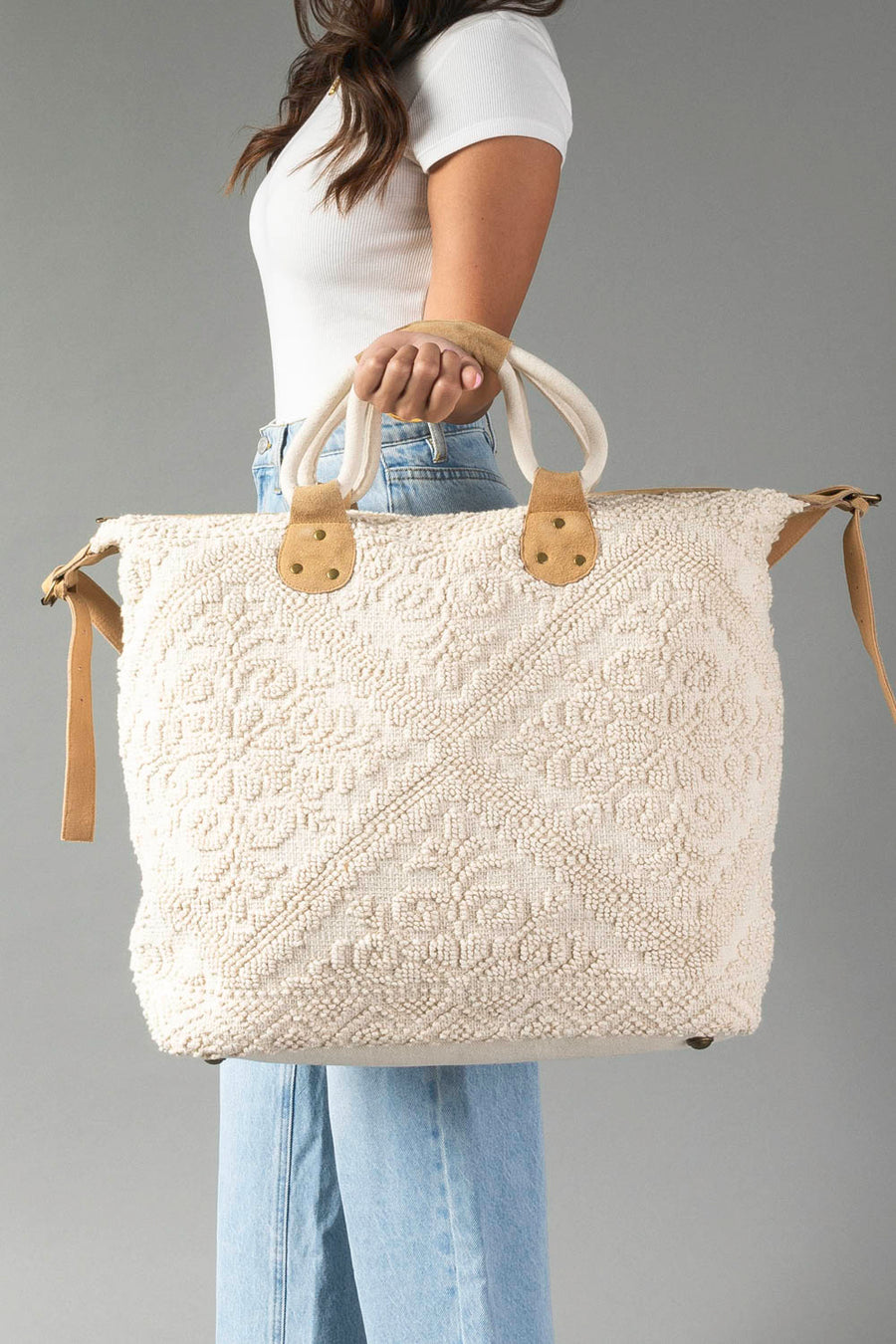 A woman in a white top and blue jeans holds the Panache Apparel Collection’s Cream Vintage Inspired Textured Tote, featuring short handles and tan accents, against a plain gray background.