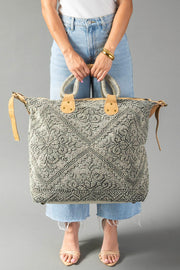 A person in blue jeans and sandals holds the Gray Vintage Inspired Textured Tote by Panache Apparel Collection, featuring suede leather handles and a zipper closure, against a plain gray background.