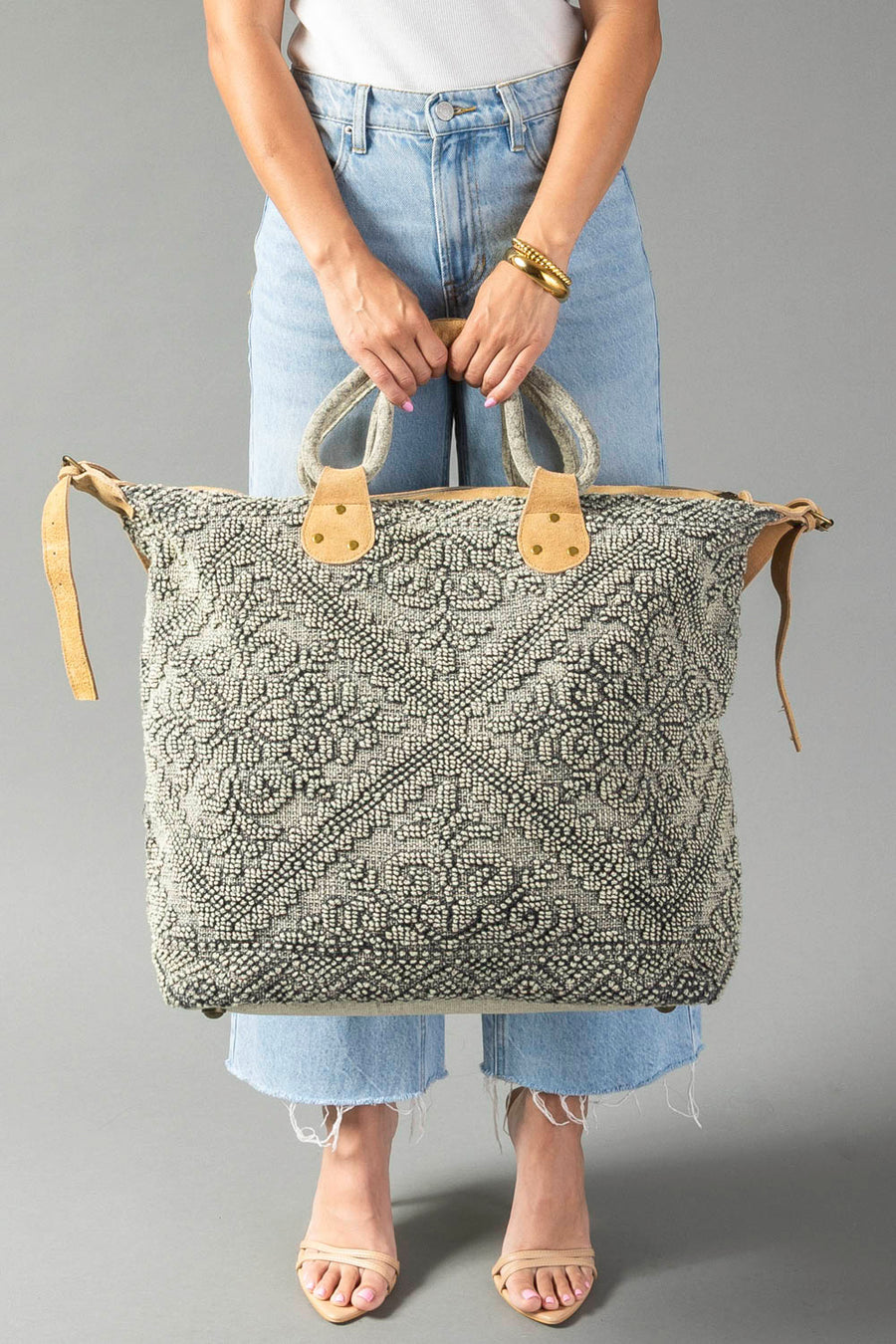 A person in blue jeans and sandals holds the Gray Vintage Inspired Textured Tote by Panache Apparel Collection, featuring suede leather handles and a zipper closure, against a plain gray background.