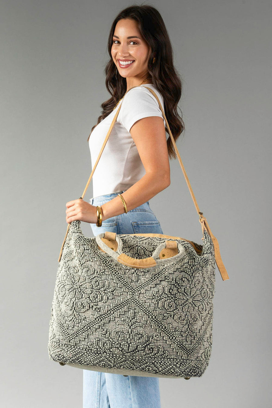 A woman smiles in a white tee and light jeans, carrying the Panache Apparel Collection Gray Vintage Inspired Textured Tote with tan suede handles and zipper closure against a plain gray background.